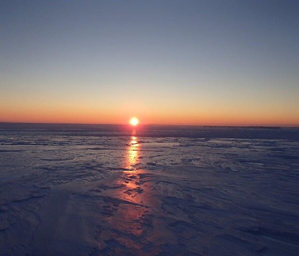 The sun sets over an icy plateau with light creating a triangular column reflecting in the slick surface