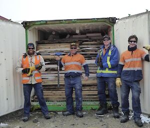 Four men with tools stand in front of an open shipping container filled with wood which once made up an old hut