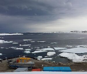 Preparation at wharf for resupply shows trucks, boats and buildings on a dirt shore with lots of sea ice in the harbour