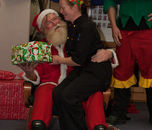 The femaile chef receives a Secret Santa gift while sitting on Santa’s lap — Santa is played by a male expeditioner with real white beard