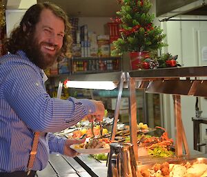 A handsome bearded male expeditioner serves himself at the buffet while smiling broadly for the camera