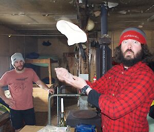 Making pizza at Wilkes, a bearded expeditioner tosses pizza dough into the air, while another male expeditioner looks on in the background