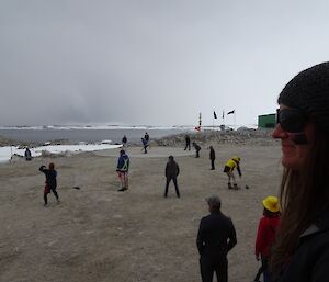 Cricket match on station with a woman smiling in the foreground, an Aussie flat on her cheek. Expeditioners play cricket with water in background.