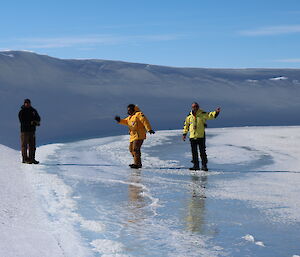 Three male expeditioners standing on ice, one waving to camera