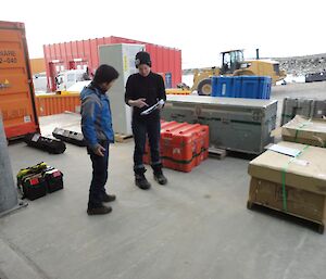 A male and female scientist are checking cargo against a list. It surrounds them on concrete outside station.