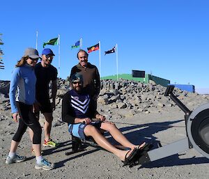 A group of four, three standing and one sitting on a rowing machine, outside at Macquarie Island station