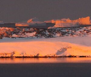 Sunset overlooking Newcomb Bay shows water in foreground and a snowy shore in the background with some rocks poking out