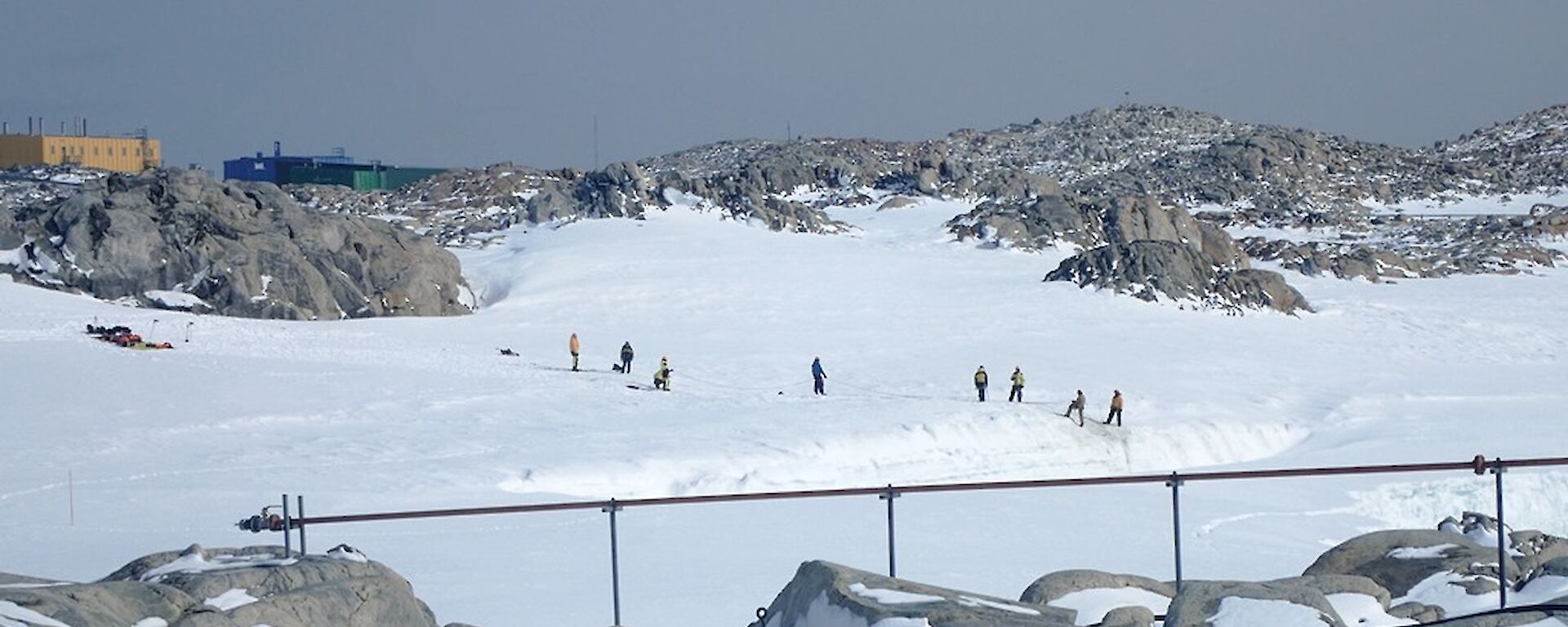 Team of expeditioners training for stretcher rescue on the edge of a crevasse with station buildings in the background.