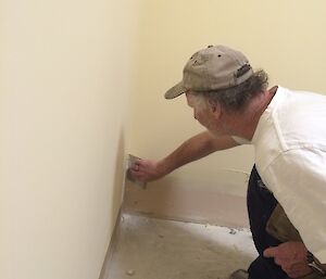 Jeff kneeling down in the corner of a room applying plaster filler with a metal spatula.