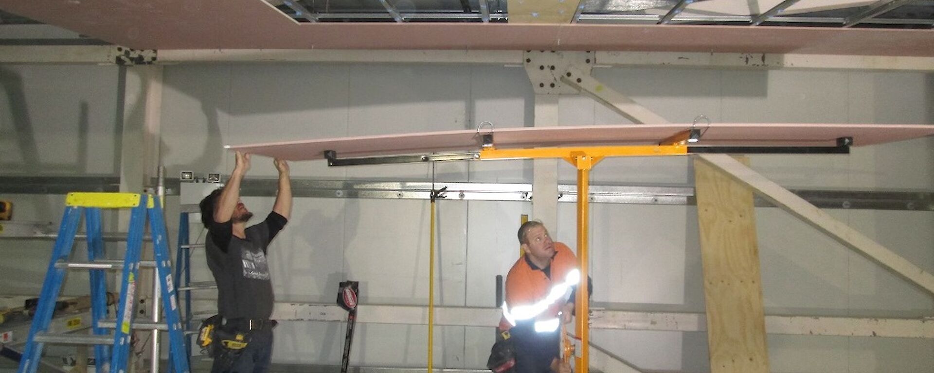 Two carpenters with a sheet of plaster on the plaster lifting device as they line it up ready for the lift it up to the ceiling supports.