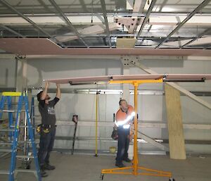 Two carpenters with a sheet of plaster on the plaster lifting device as they line it up ready for the lift it up to the ceiling supports.