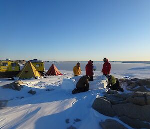 The team working on the Igloo with two polar pyramid tents and a yellow Haggland vehicle to the left side and the sea ice forming on O’Brien Bay off in the distance.