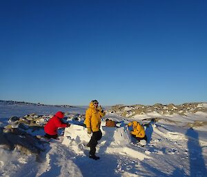 Man standing in front of igloo with rest of team working on a beautiful blue ski day.