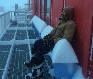 Expeditioner sitting outside building with snow on seats, in snowy conditions