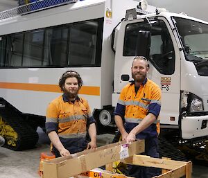 Nick and Nate getting parts out of a cardboard box to work on a big over snow bus in the background