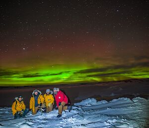 Michael, Tom, AJ & Kieran crouched together on the snow with a green aurora behind in the night sky.