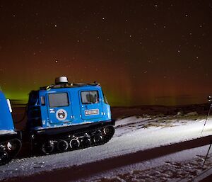 An expeditioner standing at their camera on a tripod taking a photo of the red and green aurora with a blue Hugglunds over snow vehicle in the foreground.