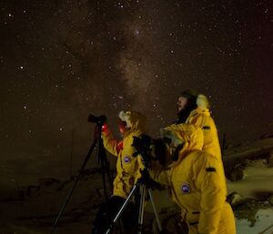 Three expeditioners standing behind cameras on tripods under star filled night sky.