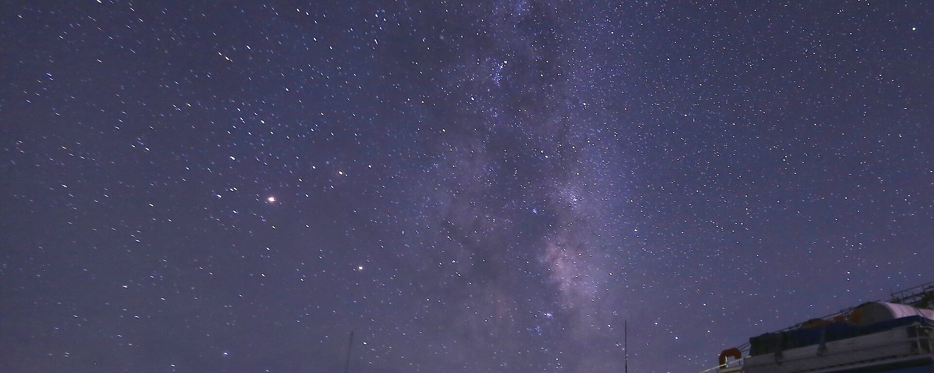 The night sky filled with stars and the Milky Way with a blue Hägglunds in the foreground on the right-hand side.