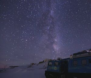 The night sky filled with stars and the Milky Way with a blue Hägglunds in the foreground on the right-hand side.