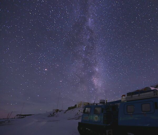 The night sky filled with stars and the Milky Way with a blue Hägglunds in the foreground on the right-hand side.