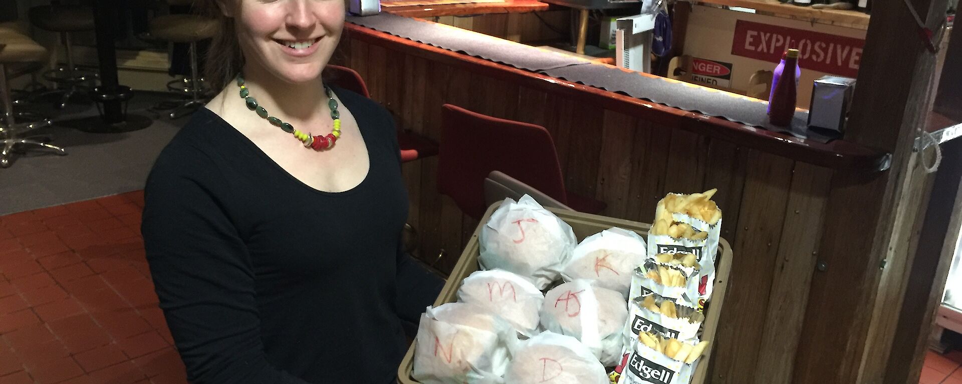 Rachel carrying a tray full of burgers and chips with the bar area in the background.