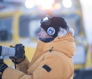 Katie watches as the plane flies overhead while holding her camera with the yellow Hagglund vehicle in the background.