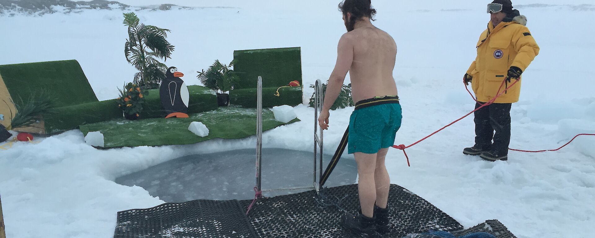 Nate with safety line attached ready to hope into the sea ice plunge pool with the fake palm trees and outdoor setting in the background.