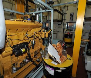 Walter (stuffed soft toy) propped up on a 200l oil drum holding a clip board and wearing ear muffs in front of a CAT engine in the main power house.