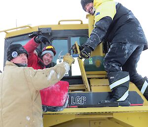 Three expeditioners pretending to find a builder’s claw hammer in the tool box of the bulldozer.