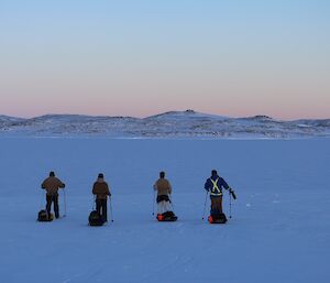 Four expeditioners in the foreground on cross country skis hauling sleds traversing away from the camera with land off in the distance.