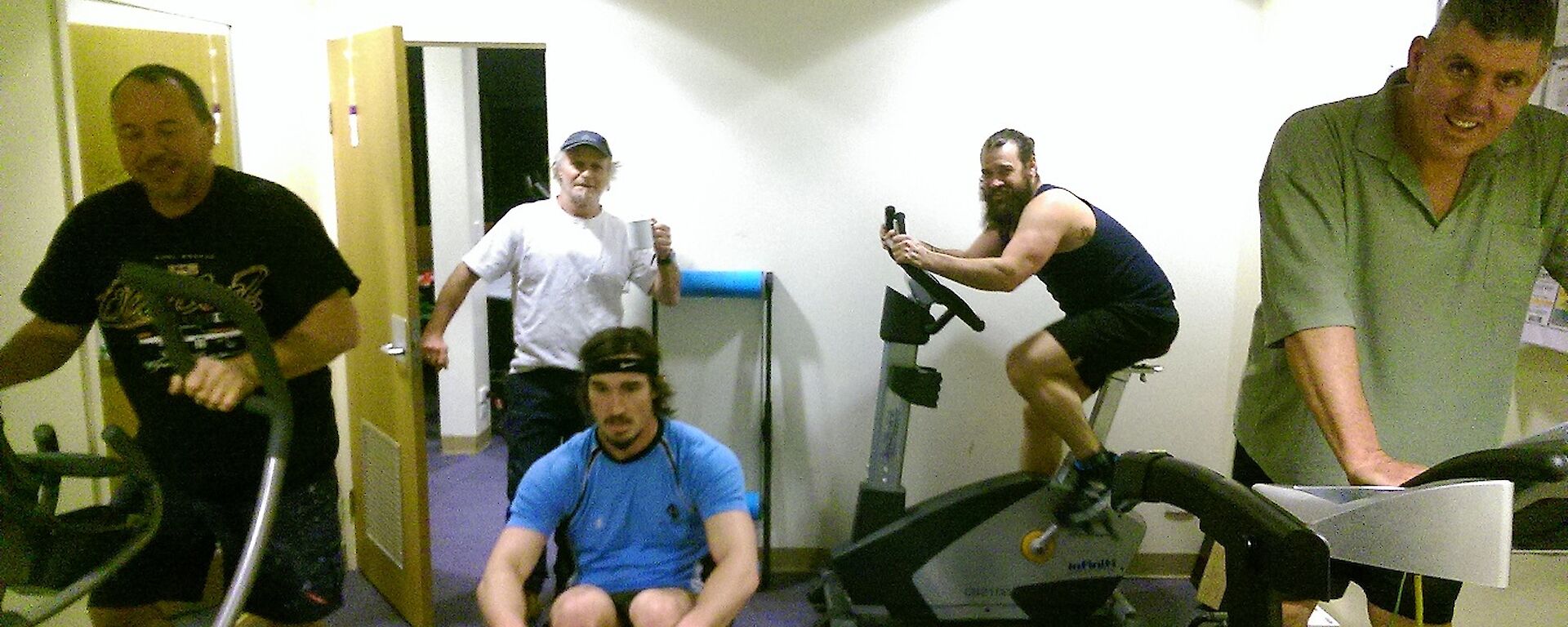 Four expeditioners exercising, one each on the cross trainer, rowing machine, treadmill and exercise bike with Jeff walking in with a coffee to see what is going on.