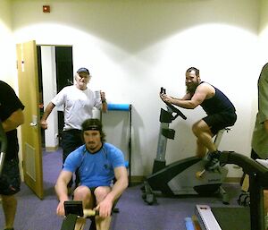 Four expeditioners exercising, one each on the cross trainer, rowing machine, treadmill and exercise bike with Jeff walking in with a coffee to see what is going on.
