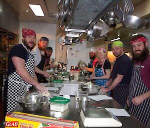 Donna the chef with six pupils in her bread cooking class lined up each side of the stainless steel work bench covered with cooking utensils.