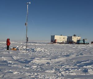 The two white sled vans and blue Hägglunds vehicle parked behind the Automated weather station where one expeditioner in a dug out hole while the other watches on from above.