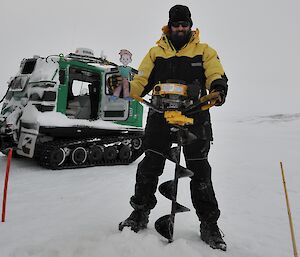 Flat Stanley being held by an expeditioner while he drills the ice to do some water sampling at O’Brien’s Bay with the green Hagglund vehicle in the background.