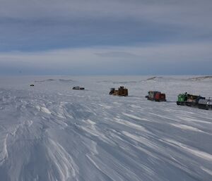 Six vehicles driving off into the distance with a thin layer of cloud cover above the stark white Plato.