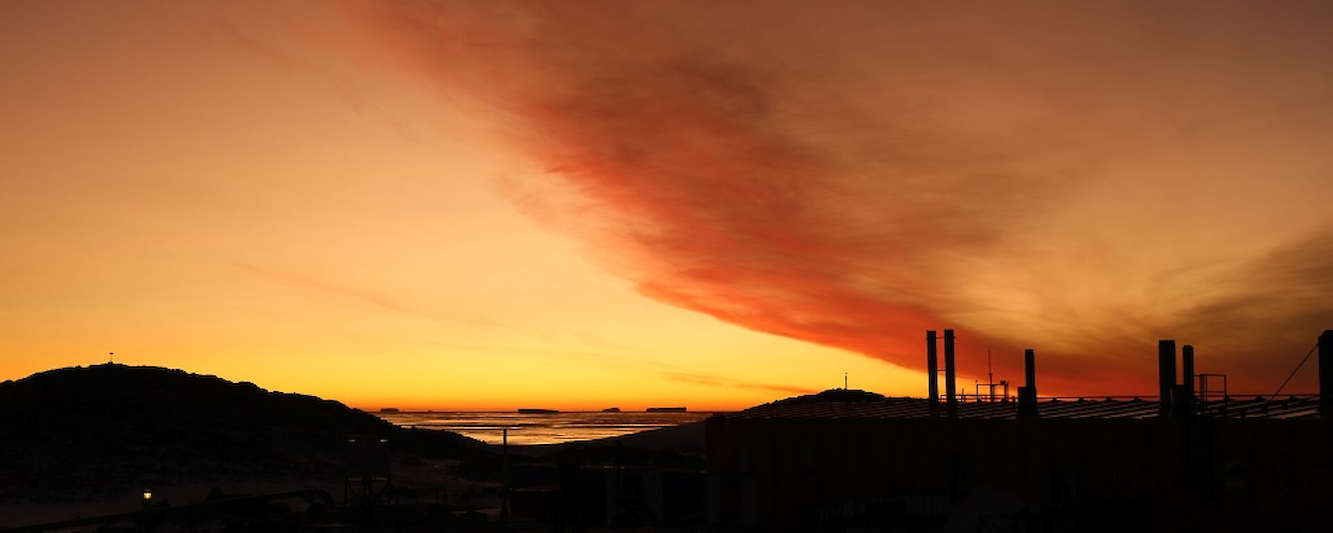 Bright orange/red sunset with the dark silhouette of hills and science building in the foreground.