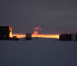 Silhouette of two buildings with the generator van and an expeditioner in the middle as the sun sets on the horizon.