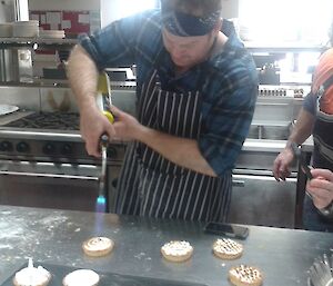 Dean using a gas torch to brulee the top of hid lemon meringue pie while AJ watches.