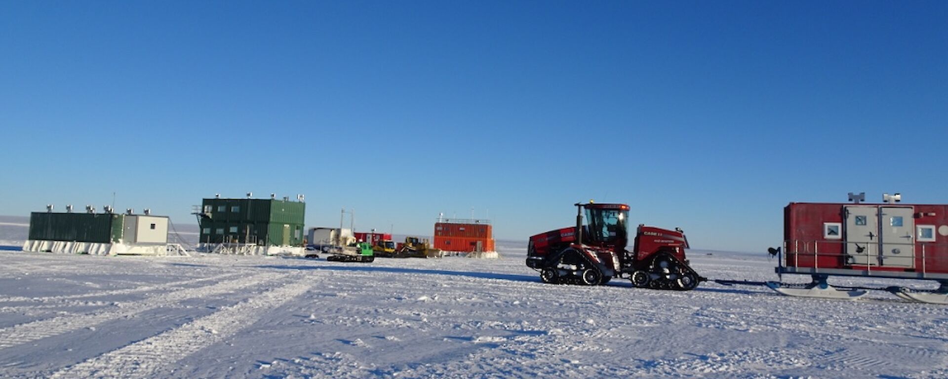 The multi-coloured builds in the background and the Quadtrak tractor towing a sled in the foreground, all on a blue sky day.