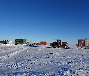 The multi-coloured builds in the background and the Quadtrak tractor towing a sled in the foreground, all on a blue sky day.