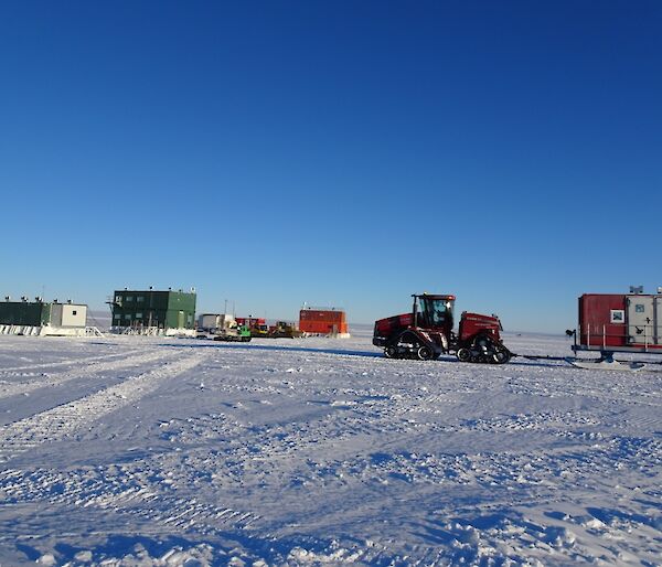 The multi-coloured builds in the background and the Quadtrak tractor towing a sled in the foreground, all on a blue sky day.
