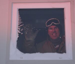 Photo taken from outside a building of an expeditioner looking through the window with a big smile and waving