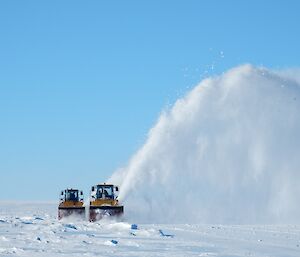 Two snow blowers operating one just behind the other with big streams of snow shooting into the air as they blow snow off the runway