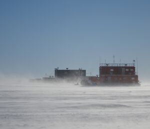 Wilkins camp building in the distance with low blowing snow on a blue sky day