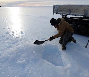 Expeditioner kneeling next to a hole in the blue ice runway that his is cleaning out with a shovel of ready for repair.