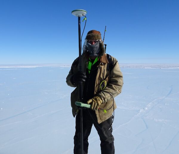 Expeditioner rugged up for the cold weather holding the survey equipment on a blue sky day.