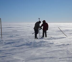 Silhouette of two expeditioners working with the survey equipment on a very cold, sunny blue sky day with the wind blowing snow along the ground.