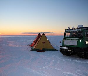 Two polar pyramid tents set up on the Plato with the green Hägglunds parked next to them as the sun goes down on the horizon.